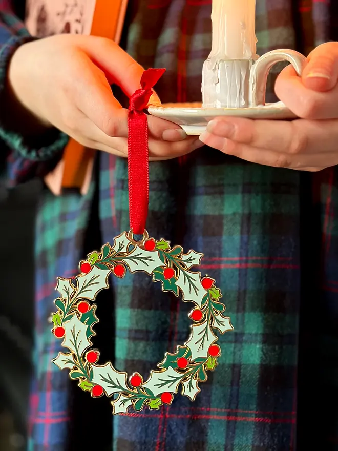 A child wearing a tartan dress holds a vintage-style enamel Christmas wreath ornament with green holly leaves and red berries, hanging from a red ribbon, alongside a small white candleholder with a lit candle.