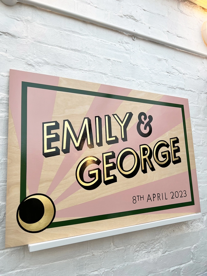 Modern wedding sign featuring the couple's names in gold leaf, outlined in black, and the wedding date, with a sunburst background and a border. Shown against a white brick wall, at an angle. 