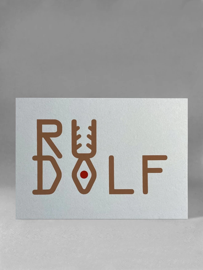 Rudolf is spelt out in brown ink, stacked to look like a reindeer - with a red dot depicting the nose. The grey card is stood in a light grey studio set, front on.