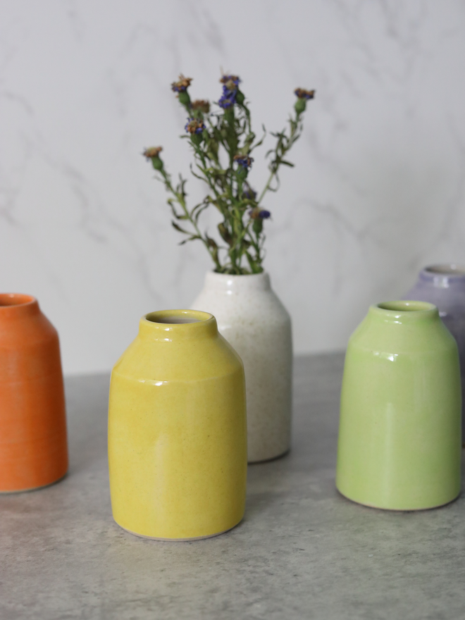 A close-up of five handmade ceramic bud vases grouped together on a grey surface, in orange, yellow, speckled white, spring green, and lavender glazes, with dried purple wildflowers in the white vase, against a marble background