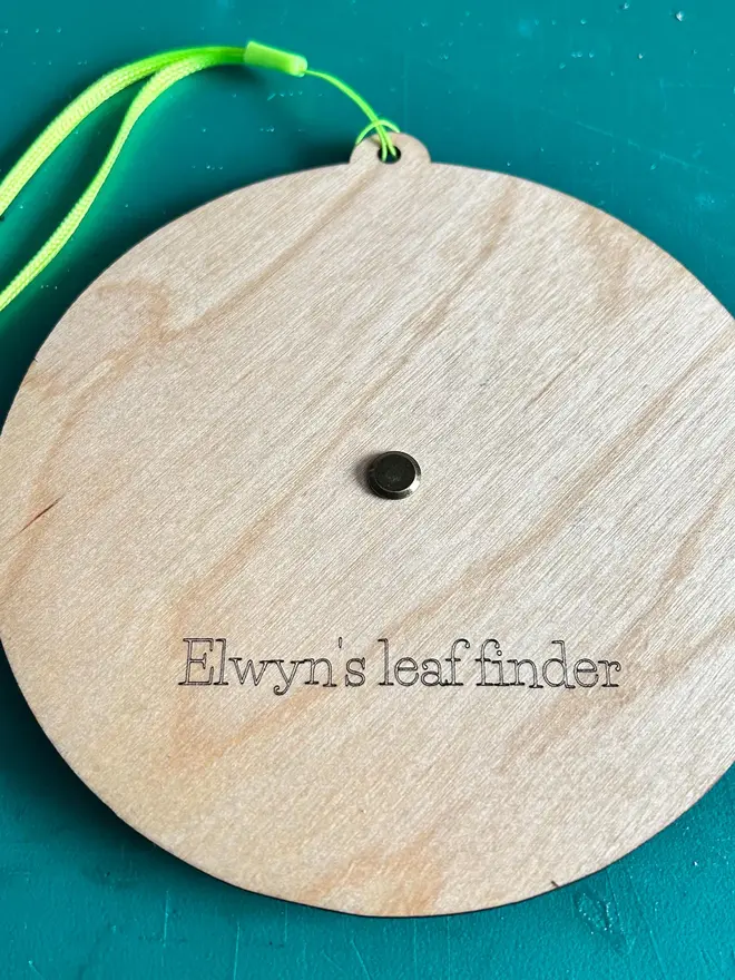 Back of the round wooden leaf identification dial engraved with detailed leaf illustrations and rotating pointer on a blue table. 