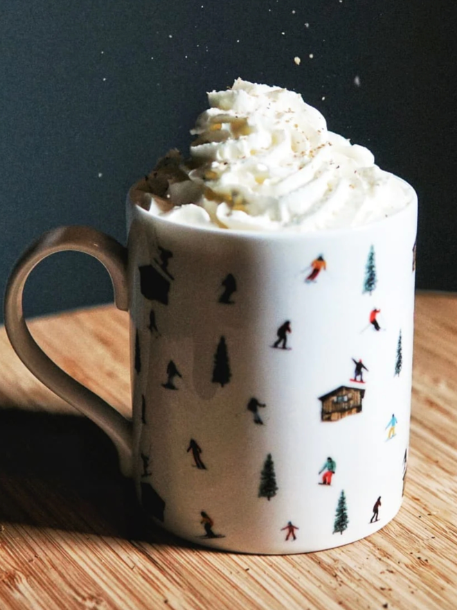 Ski Hut Mug. A white mug set on a white table while in use holding a hot drink. The design is a pattern of multicoloured skiers, ski huts, and trees.