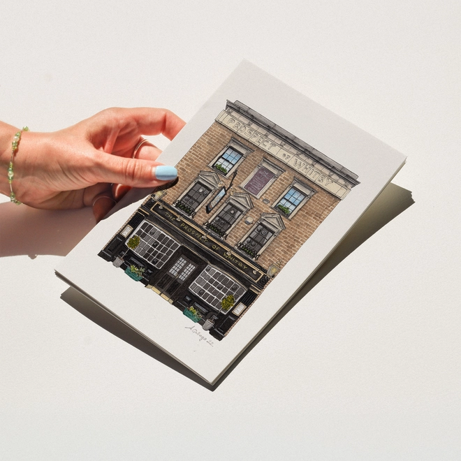 A hand with blue nail polish holding a print featuring an illustration of the Prospect of Whitby pub against a white background.