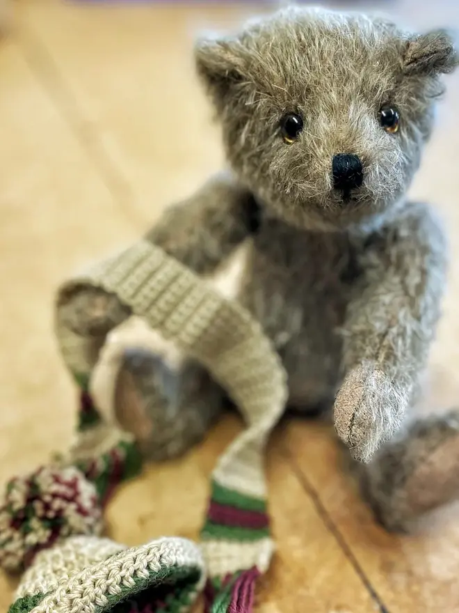 Brown teddy bear sitting upright wearing a knitted beige hat and scarf with green and burgundy details on a wooden table. 