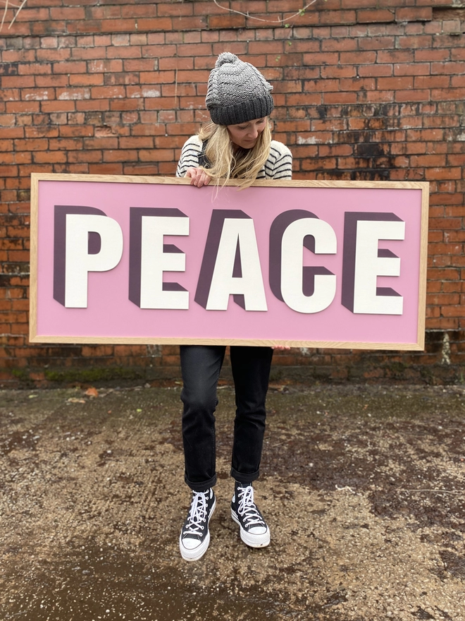 A person holding a painted wooden sign reading PEACE, with a pink background, standing in front of a brick wall