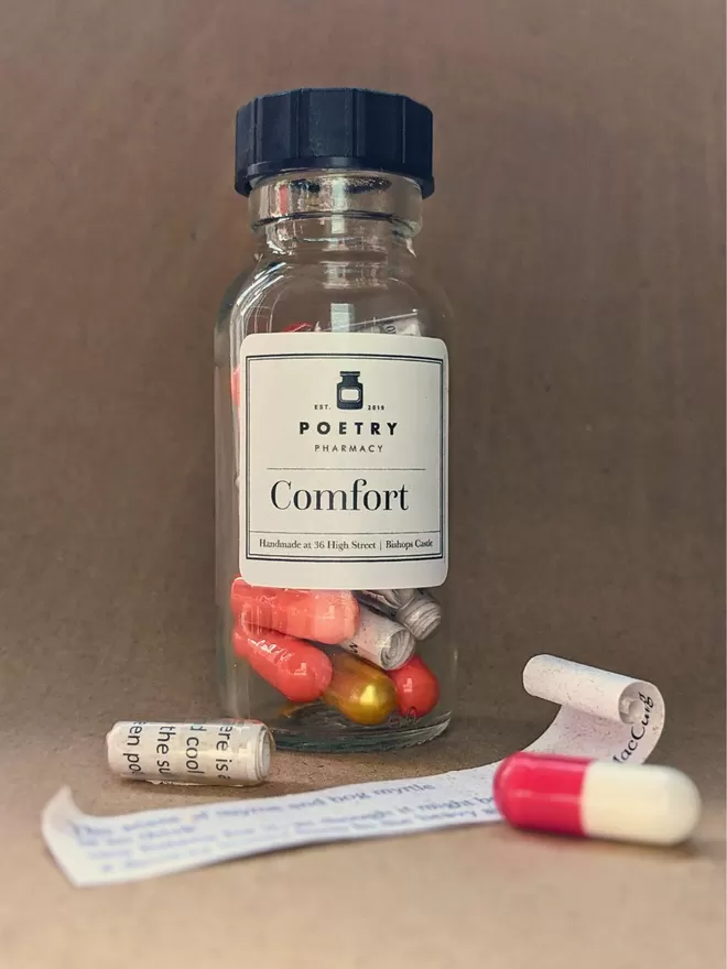 A bottle of comfort poetry pills with a quote on a slip of paper and a clear capsule with a rolled up quote inside it