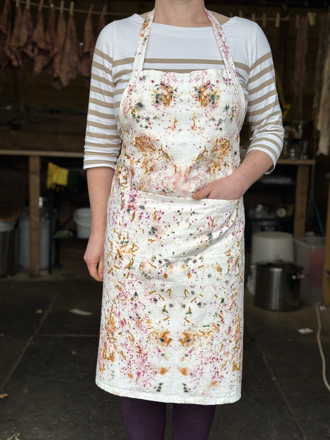  A vibrant, naturally dyed organic cotton apron with a bright, playful pattern is worn by the maker, standing outdoors in front of a cosy studio. The colours in the apron pop against the natural background, highlighting the creative and eco-friendly design. The apron’s large front pocket make it both stylish and functional, perfect for crafting or gardening. The joyful pattern reflects a love for sustainable, handmade fashion.