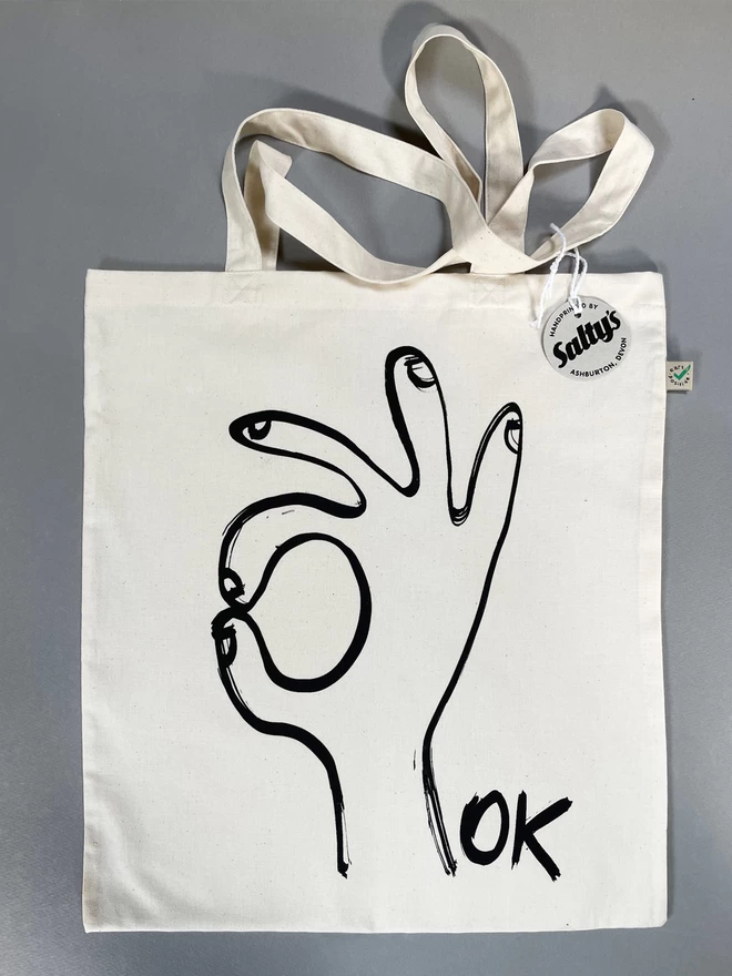 Flat lay of Ok tote bag with black ink hand drawn in an OK gesture - Saltys swing tag tied to one handle and showing the fairtrade label.