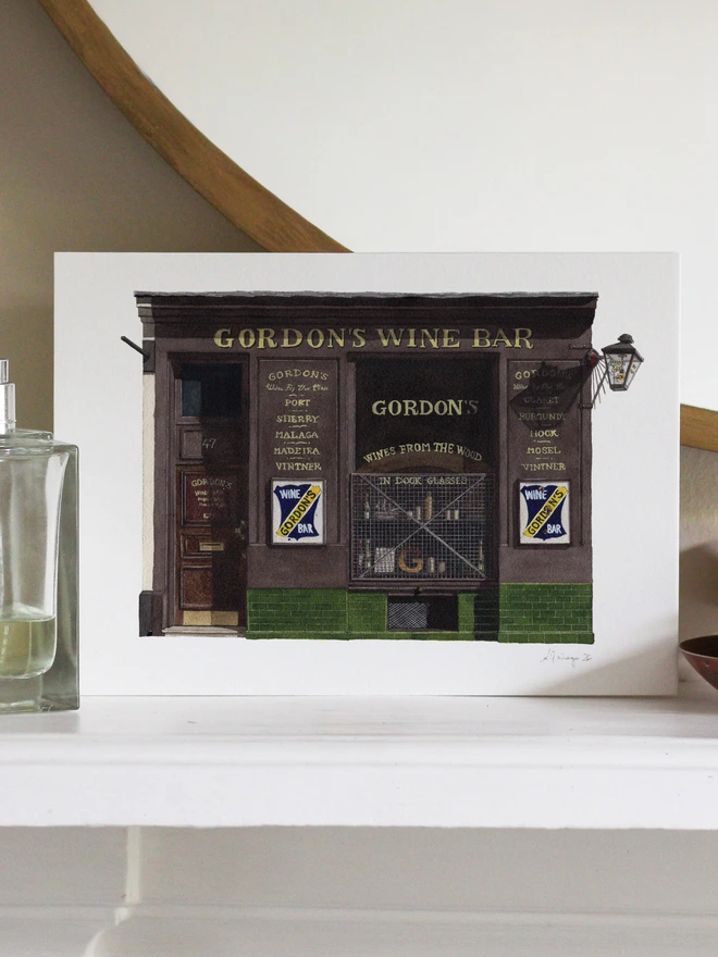 A watercolor print of Gordon's Wine Bar resting on a white shelf next to a glass perfume bottle.