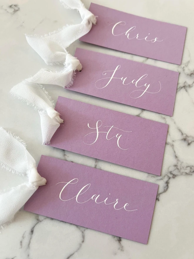 Personalised Calligraphy Wedding Lilac Place Cards, group of lilac place cards with white ribbon. 