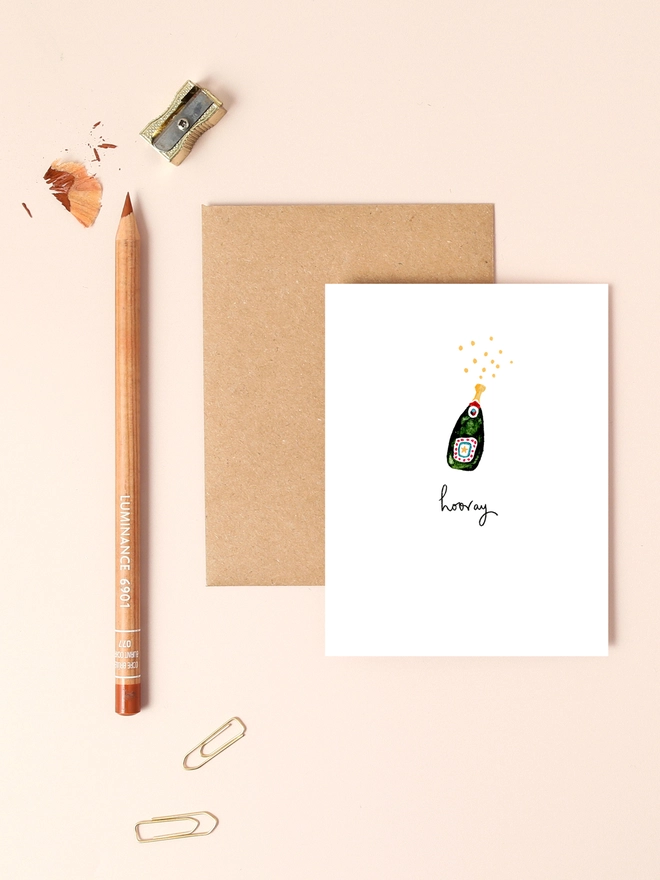 An image of a mini, rectangular white greetings card with an illustration of a champagne bottle with tiny bubbles coming out of it with a hand written 'hooray' message seen under the central illustration. The mini greetings card is placed on top of a rectangular brown kraft envelope.