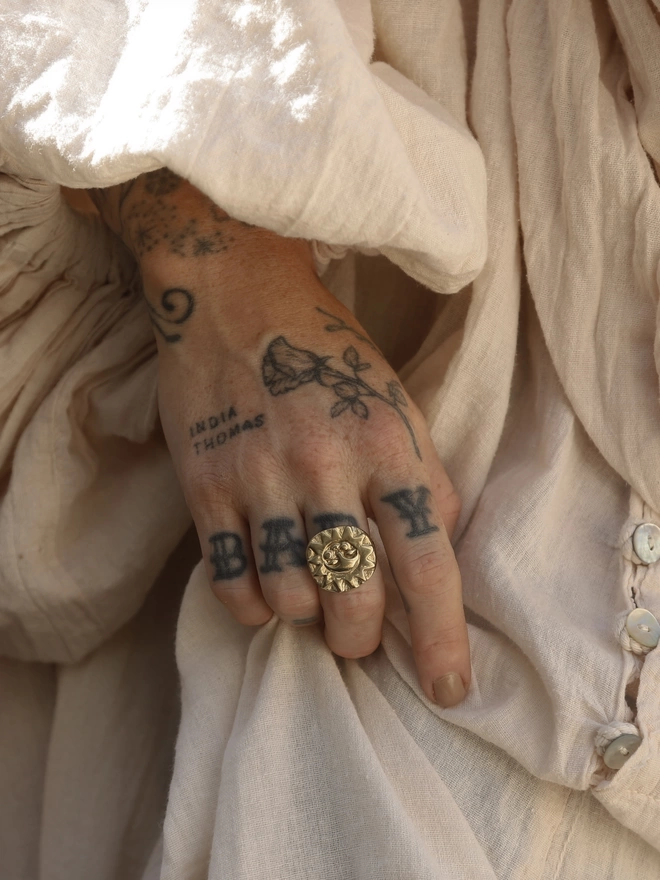 An image of a tattooed female hand wearing a gold toned brass hand carve sun face ring, with a loose and layered cream linen style fabric dress
