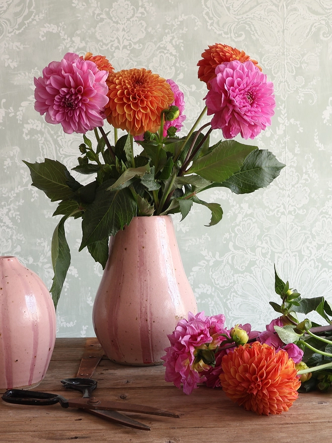 A Handmade Ceramic Stoneware Large Vase in Pink Stripe filled with a bouquet of pink and orange flowers. A medium, rounded vase in the same style sits next to the larger vase, along with additional flower stems and a pair of scissors. 