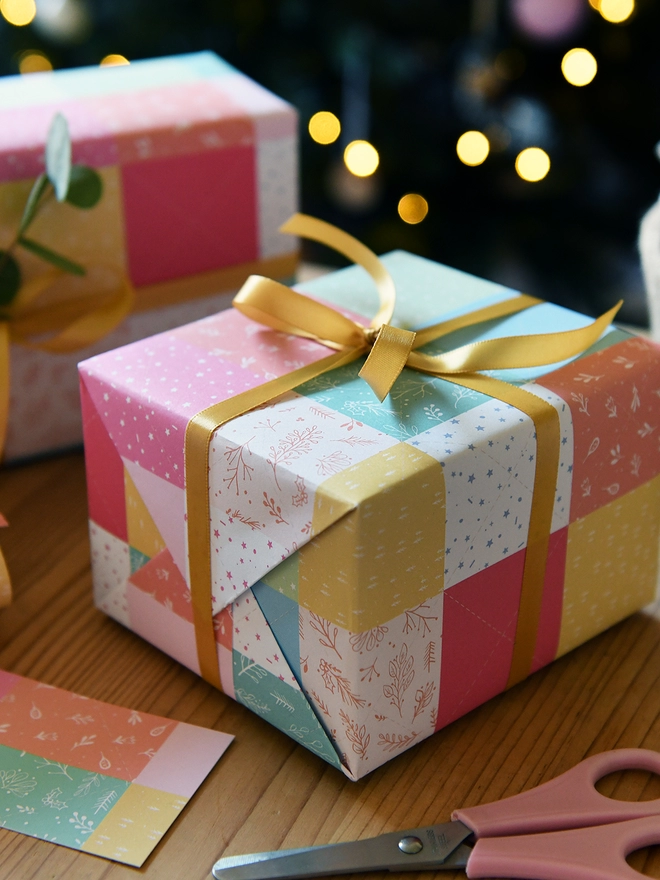 Gifts wrapped in gift wrap with a design of pastel patchwork quilt design is on a wooden table in front of a Christmas Tree.
