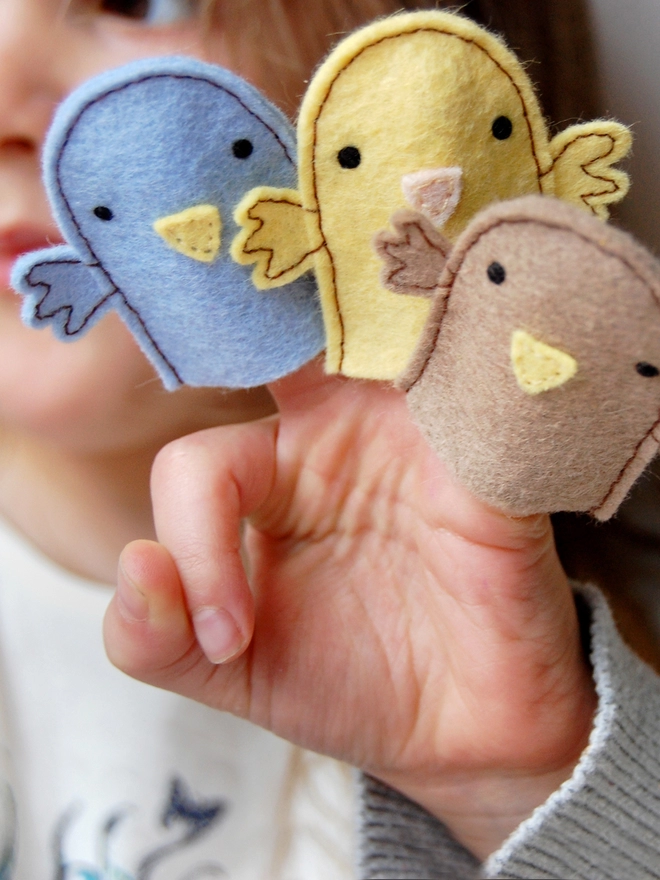 A young child is holding three homemade felt bird finger puppets in her hands. There is one yellow, one brown, and one blue bird.