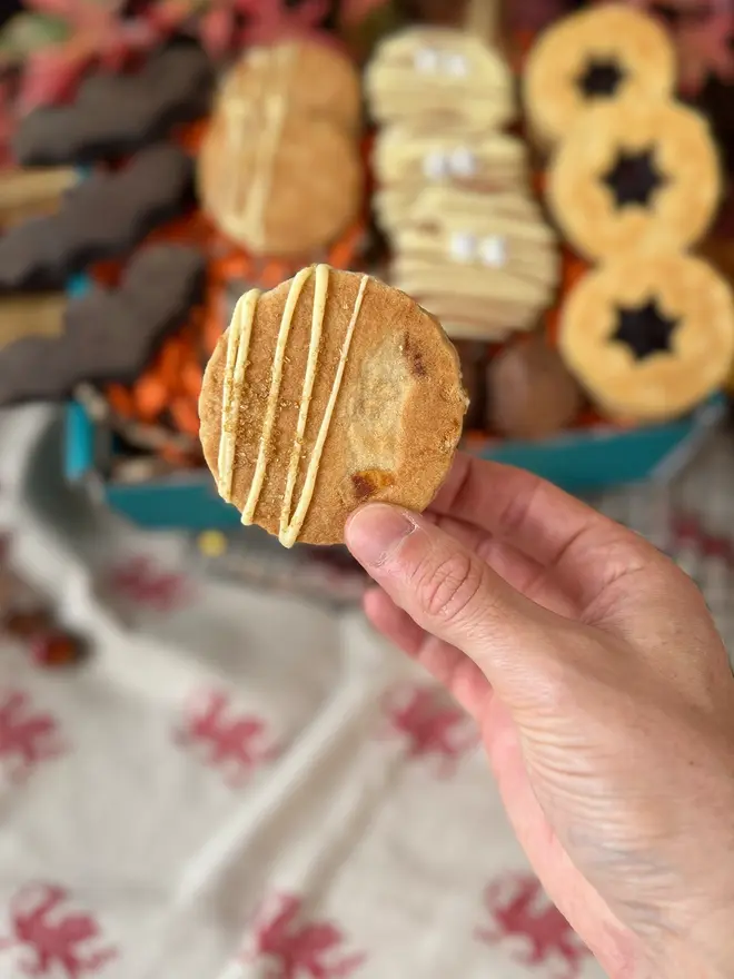 A hand holding a round golden-brown welshcake with diagonal drizzle of white chocolate icing, with assorted welshcakes in the background inside a gift box.