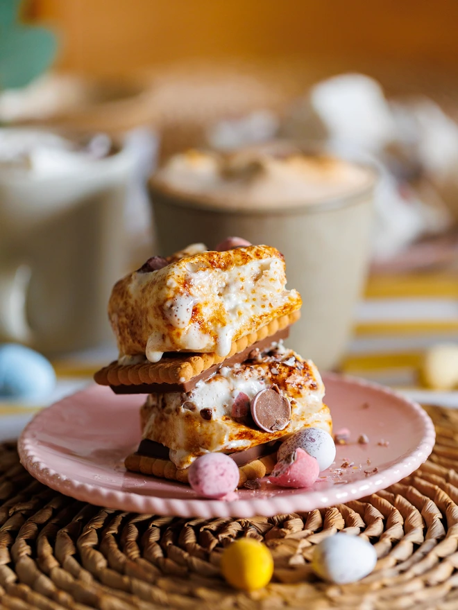 The Mallow Makers Easter Luxury S'mores and Hot Chocolate Kit Toasted marshmallows and mini eggs stacked between biscuits on top of a pink plate on a wicker place mat