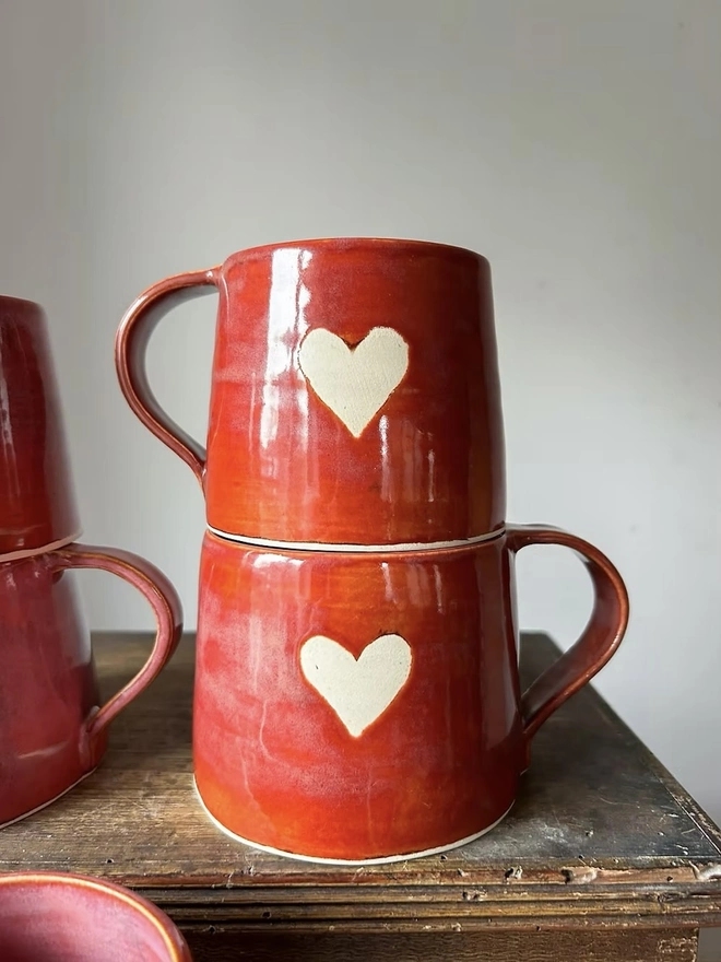 Heart Mug Cinnamon , a pair of heart mugs stacked on top of each other on a wooden surface in front of a plain surface. 