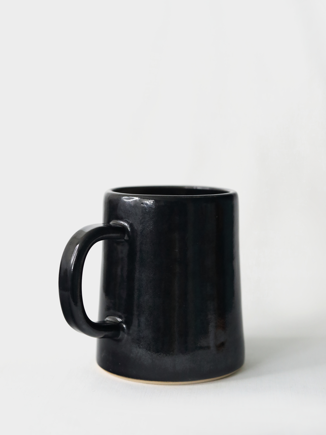 A handmade ceramic pint mug in jet black glaze with a chunky loop handle, photographed against a plain light grey background