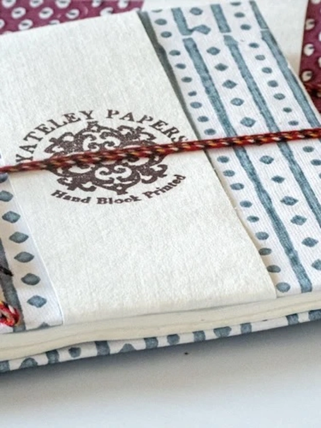 A5 Small Landscape Travel Watercolour Book. Close-up of a Yateley Papers hand block printed notebook with a slate blue stripe and diamond-dot cover, tied with red braided cord. The cream branded paper band and textured handmade paper give the stationery an artisan, giftable finish.