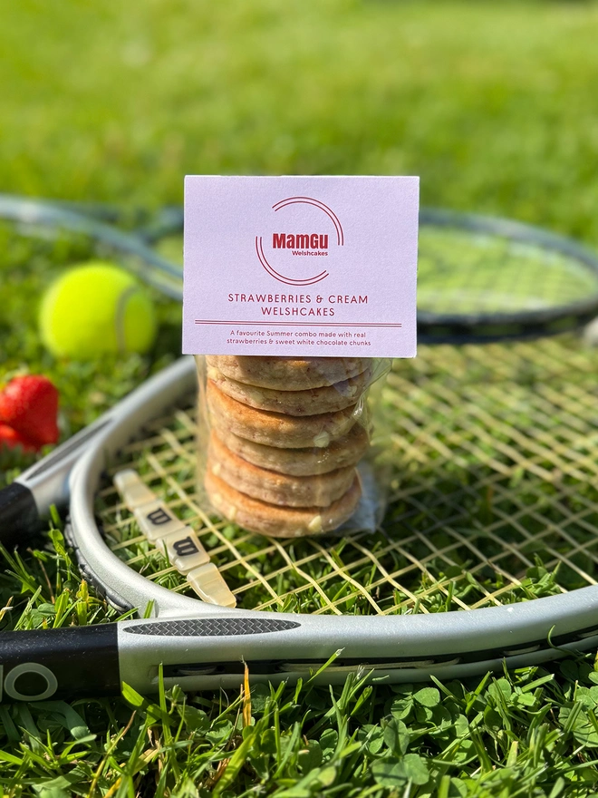 6 Pack of Strawberries & Cream Welsh Cakes with a pink MamGu label on top, sitting on tennis rackets on grass.