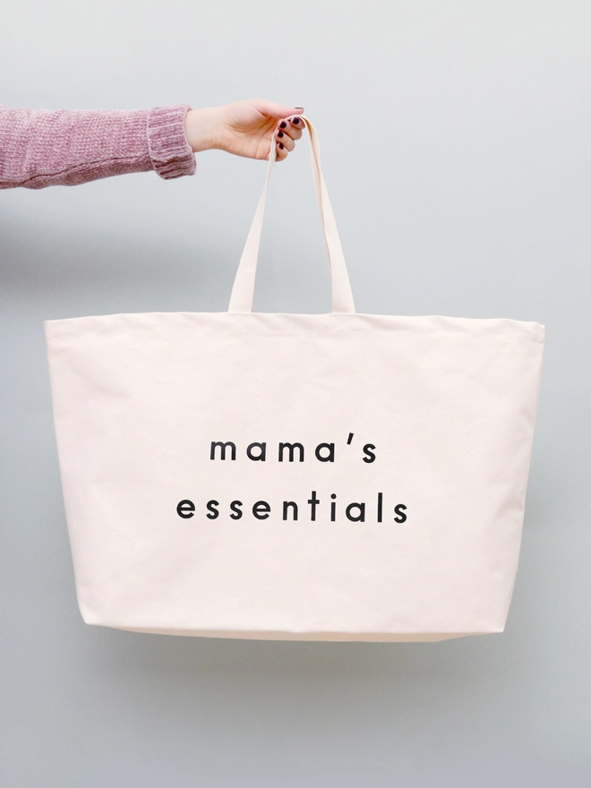 model holding out an oversized natural canvas tote bag with mama's essentials slogan