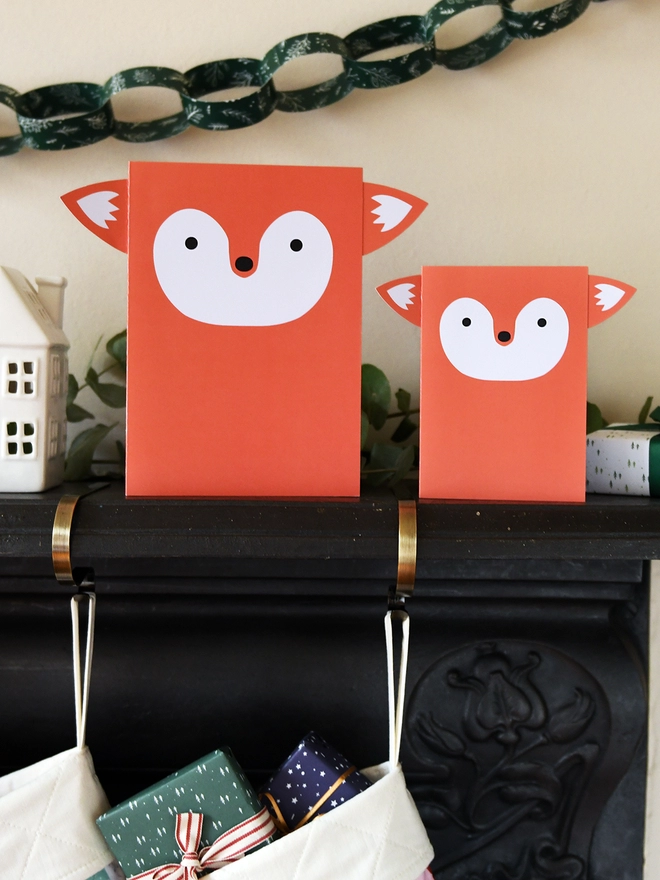 Two orange fox greetings cards stand on a black mantlepiece where two patchwork stockings are hanging from.