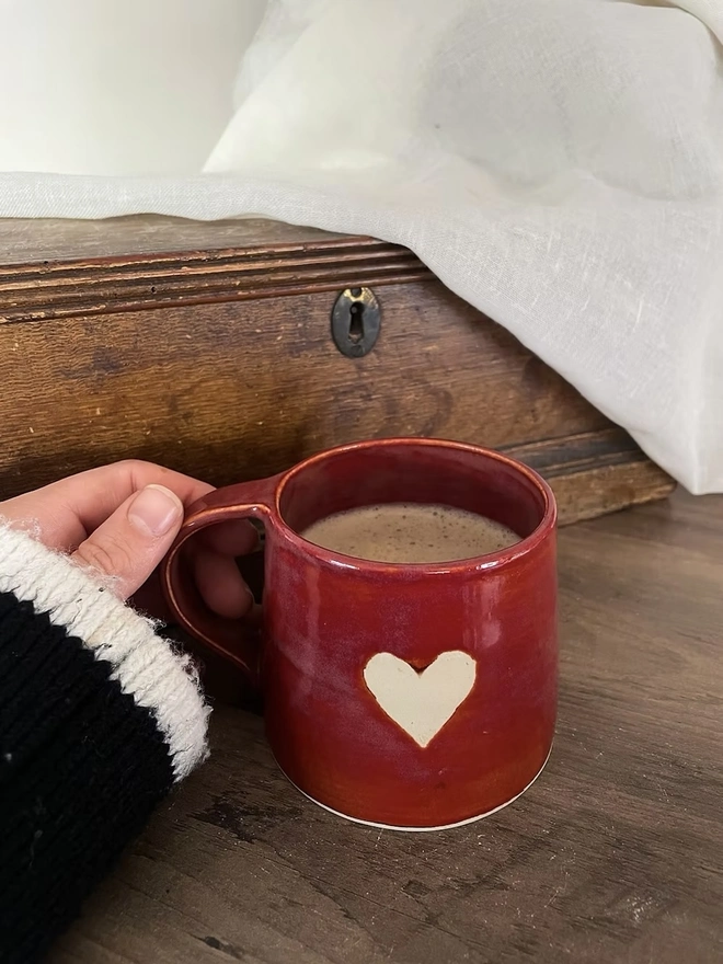 Heart Mug Cinnamon, a heart mug filled with coffee on a wooden surface being held by someone. 