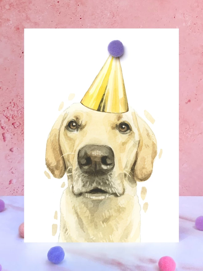 A greeting card featuring a hand painted design of a yellow labrador, stood upright on a marble surface surrounded by pompoms. 