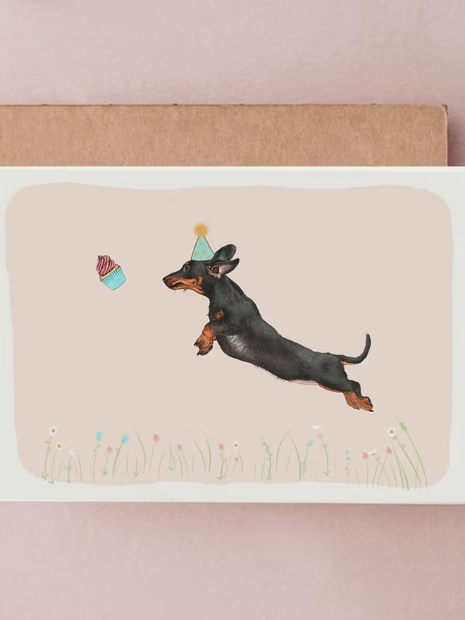 illustrated dachshund birthday card. a black and tan sausage dog jumping for a cupcake.