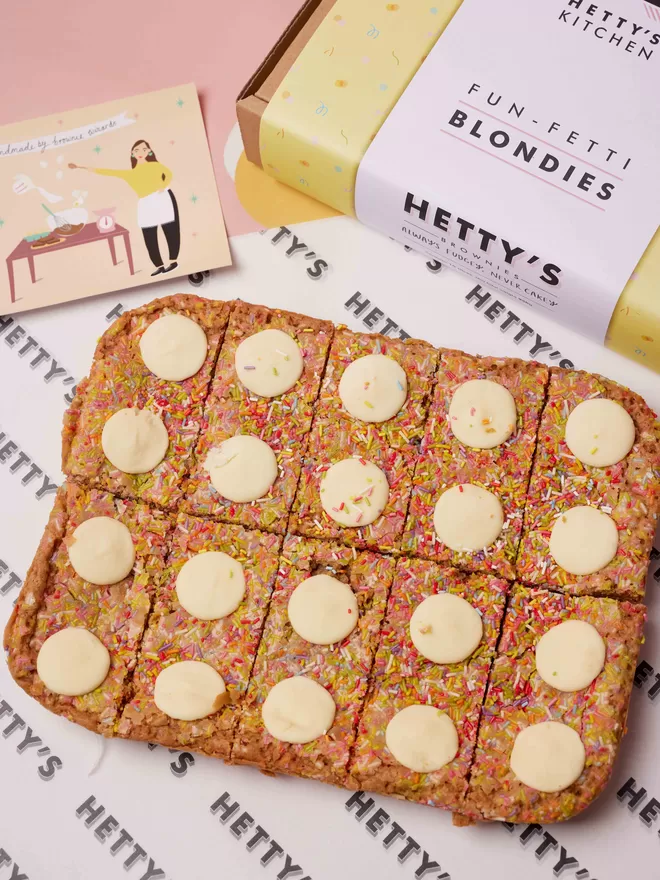 Ten slice Funfetti blondies with sprinkles and white chocolate buttons alongside branded box and postcard