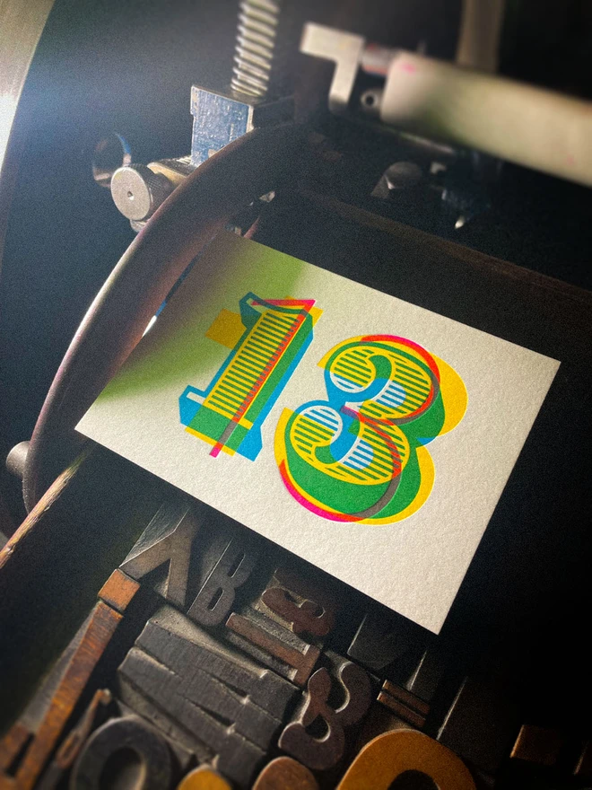 Finished handmade 13th birthday letterpress card resting on the vintage printing press, featuring a bold number 13 built from overlapping cyan, magenta and yellow inks with vibrant secondary colours where the inks overlap.
