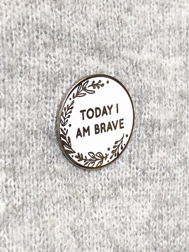 A round white enamel pin with a floral design and the words "Today I Am Brave" is pinned to grey fabric.