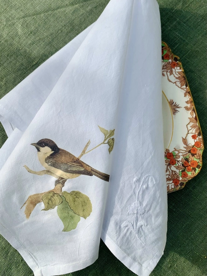 A white cotton napkin printed with a vintage illustration of a sparrow, on a vintage side plate and green linen tablecloth
