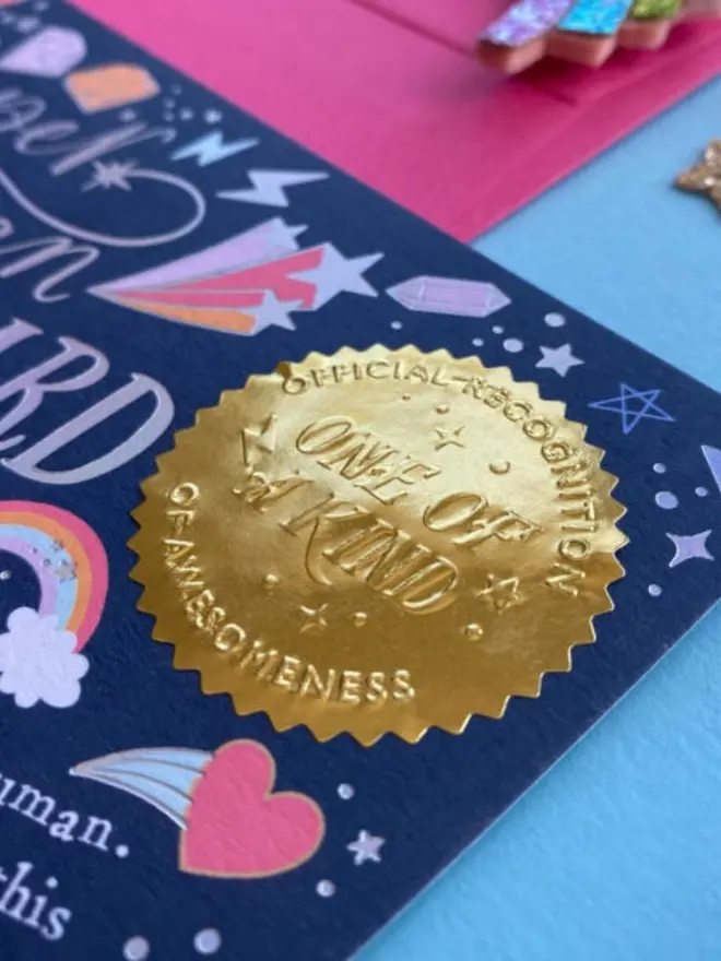 Close-up of the embossed gold "Official Recognition of Awesomeness, One of a Kind" seal. Colourful rainbow, heart and gem illustrations surround it on the navy certificate.