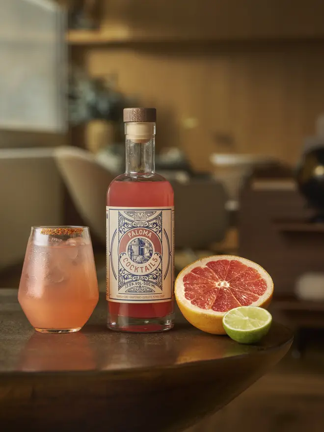 Tommy's Fizz Paloma Cocktail, a pink Paloma cocktail with a chili-salt rim and a lime wedge next to its bottle.