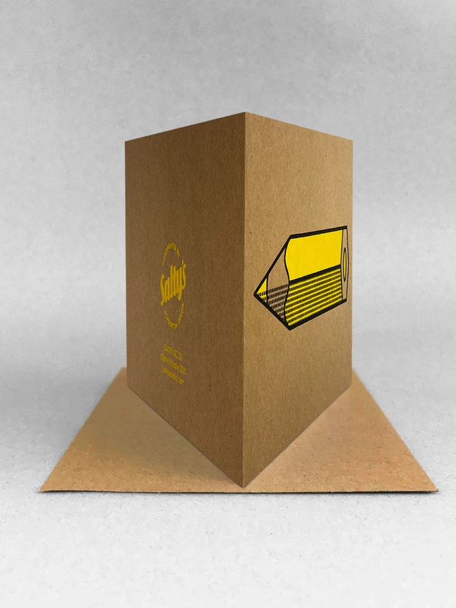 Kraft brown card, stood in a light grey studio background, also stood on a brown envelope with the fold of the card towards the camera. The yellow pencil design can be seen on the right disappearing into perspective, the left side shows the rear of the card logo and blurb. 