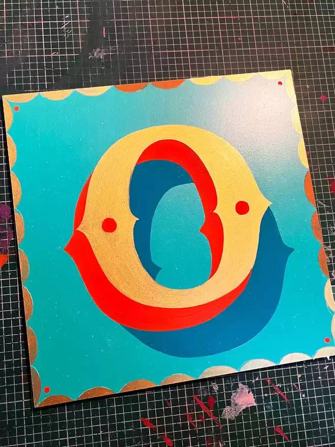 'O' painted on a turquoise background in metallic gold paint with orange shade and details, with a golden scalloped border.