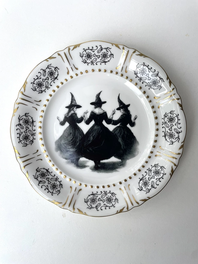 a coven of 3 witches dance hand in hand, the image is victorian in black and white and is printed directly onto an ornate vintage plate
