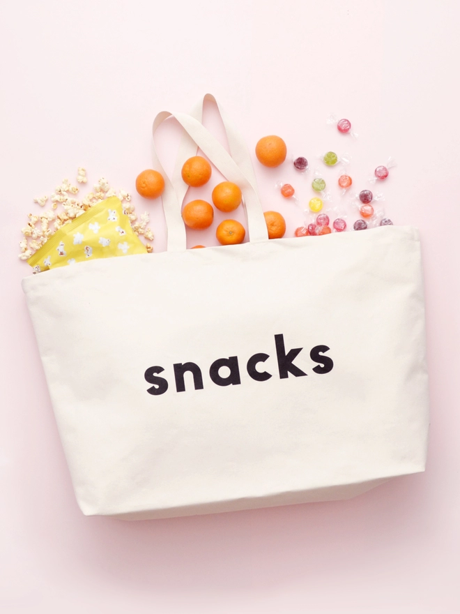 oversized natural canvas tote bag laying on a pink background with snacks spilling out