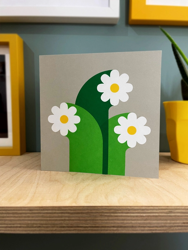 Three white flowers with oversized stems screenprinted on grey square card, Stood on a plywood shelf with picture frames around the edges.