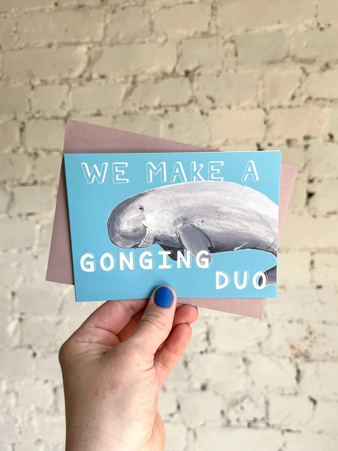 a bright blue greetings card featuring an illustration of a dugong with the text “we make a gonging duo” with a muted purple envelope