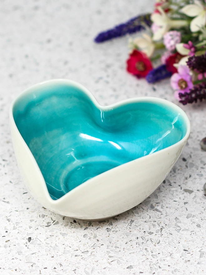 porcelain turquoise heart shaped bowl. a white heart shaped bowl glazed turquoise on the inside. photographed on a table with a bunch of flowers in the background.