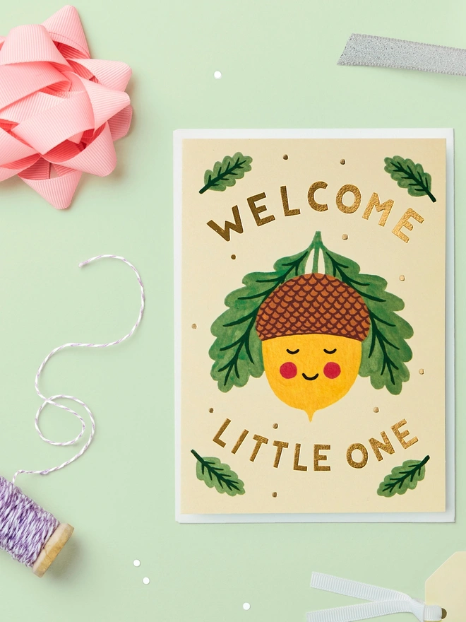 New baby card with an acorn illustration on it paired with a white envelope on a green background with confetti & ribbons scattered around.