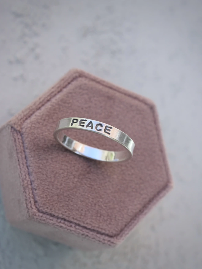 A sterling silver ring band with the word 'peace' stamped on it, resting on a hexagonal pink velvet box against a grey-green background.