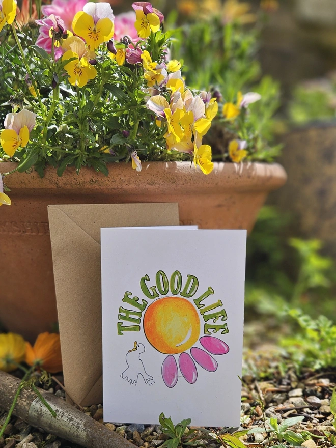 A white card with an illustration of a pink flower and a white bird with The Good Life written using a retro style font in green.