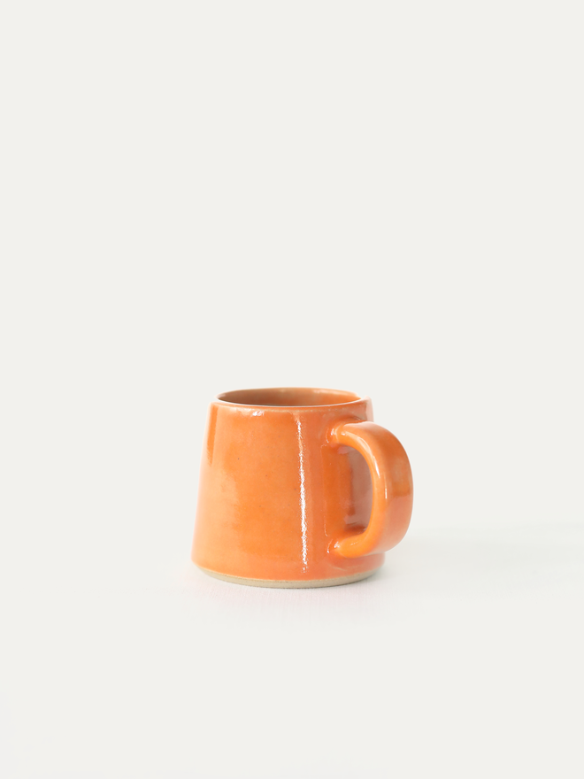 A handmade ceramic espresso mug in mandarin orange glaze, with a chunky loop handle, photographed against a plain light grey background