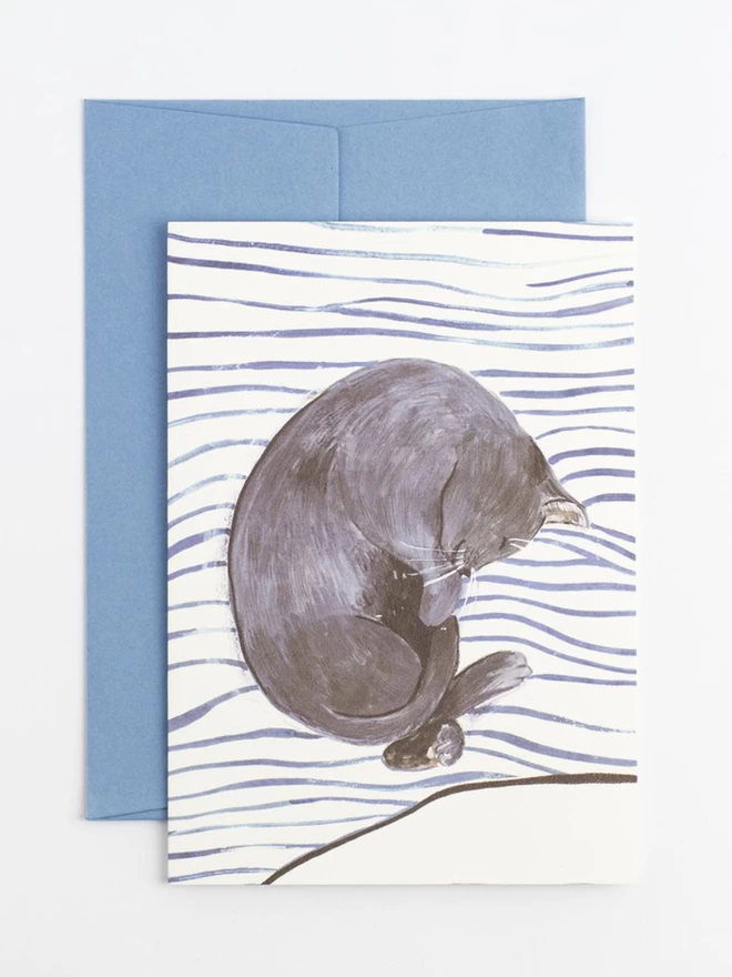 A grey cat curled up asleep on top of striped bedding with corresponding blue envelope