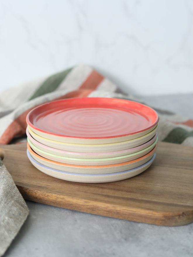 A stack of six handmade ceramic cake plates on a wooden board, topped with a cherry red plate showing a concentric swirl texture, with coloured rim edges visible beneath in yellow, blush pink, spring green, mandarin orange, and lavender, styled with a striped linen cloth against a marble background