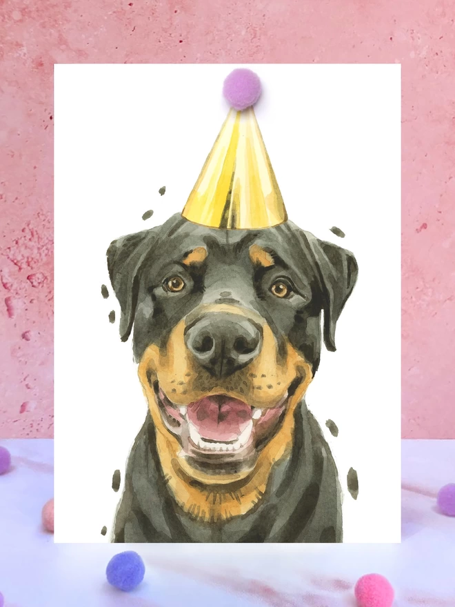 A greeting card featuring a hand painted design of a rottweiler, stood upright on a marble surface surrounded by pompoms. 